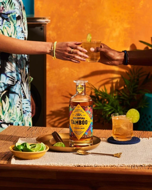 Two individuals pass a cocktail drink over a wooden counter with a bottle of rum, limes, and a gold spoon. Warm orange background.