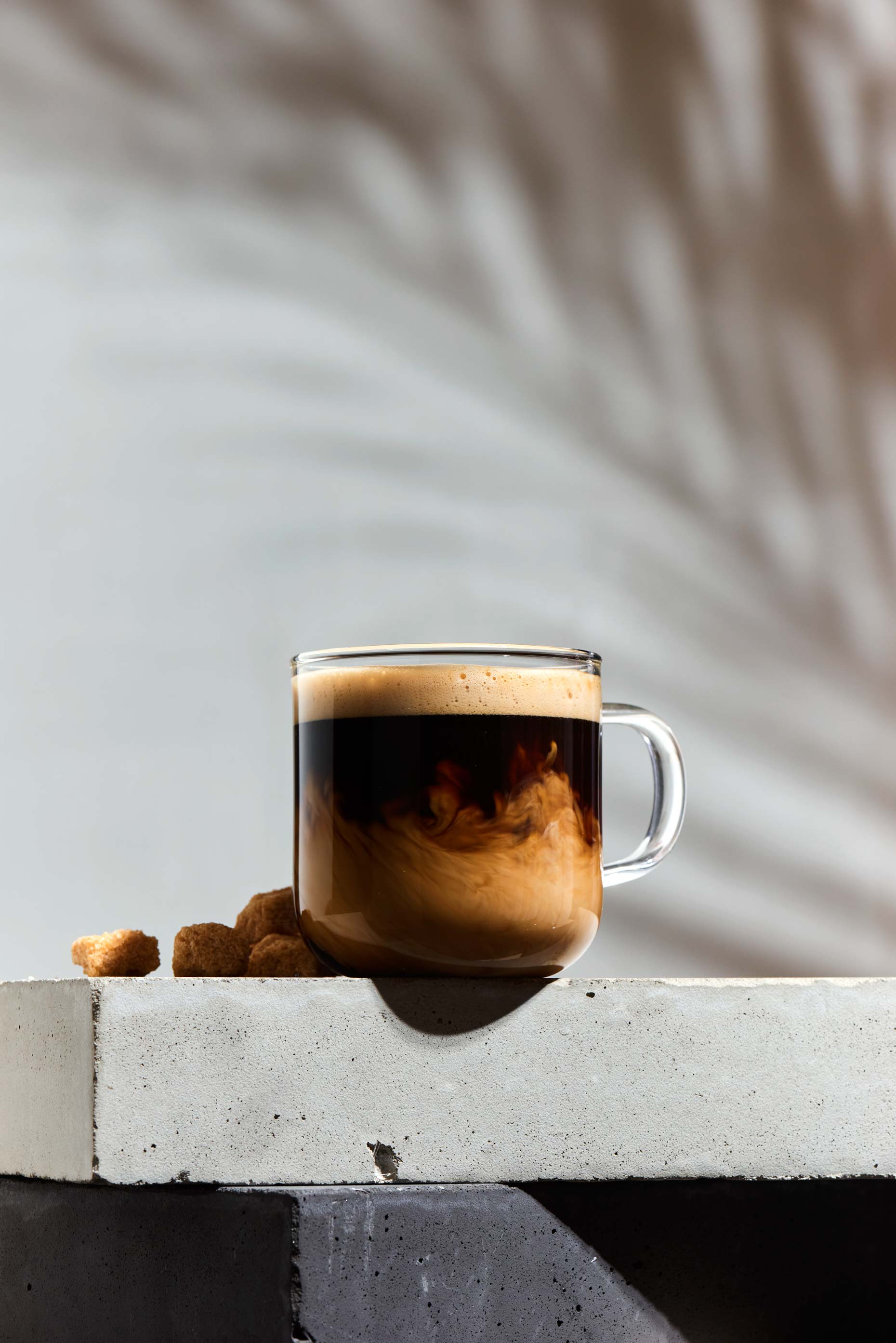 Clear glass mug with coffee and cream on a concrete surface, with brown sugar cubes, against a blurred light background with a shadow.