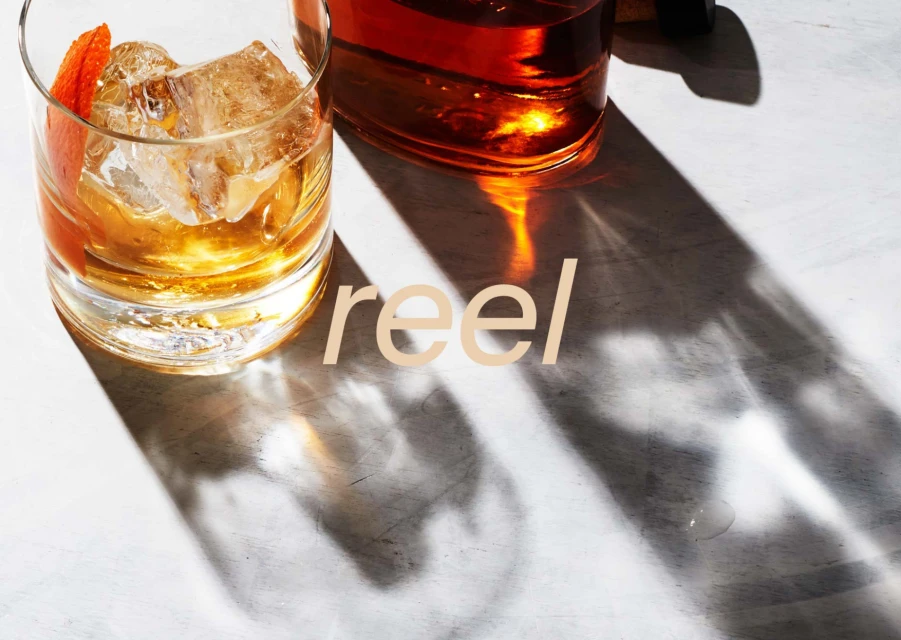 A cocktail with ice and an orange garnish sits on a white surface next to a bottle, casting sharp shadows, with 'reel' overlaid.