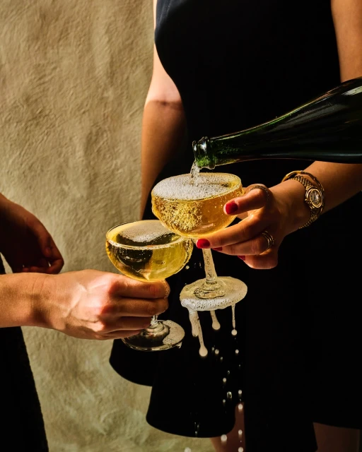 Two women pouring sparkling wine into coupe glasses with a textured background, featuring golden liquid and dark dresses.