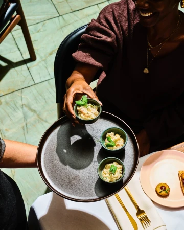 A server holding a dark tray with small bowls of food, while a smiling seated diner looks on at a table.
