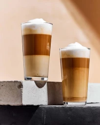 Two tall layered latte drinks in clear glasses with foam on concrete blocks against a warm neutral background.