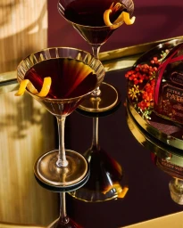 Two elegant cocktail glasses filled with dark amber liquid and orange twist garnishes on a reflective dark surface.