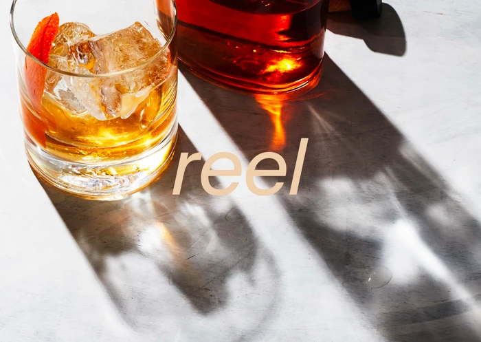 A glass of amber whiskey with ice and an orange twist next to a decanter on a light marble surface.