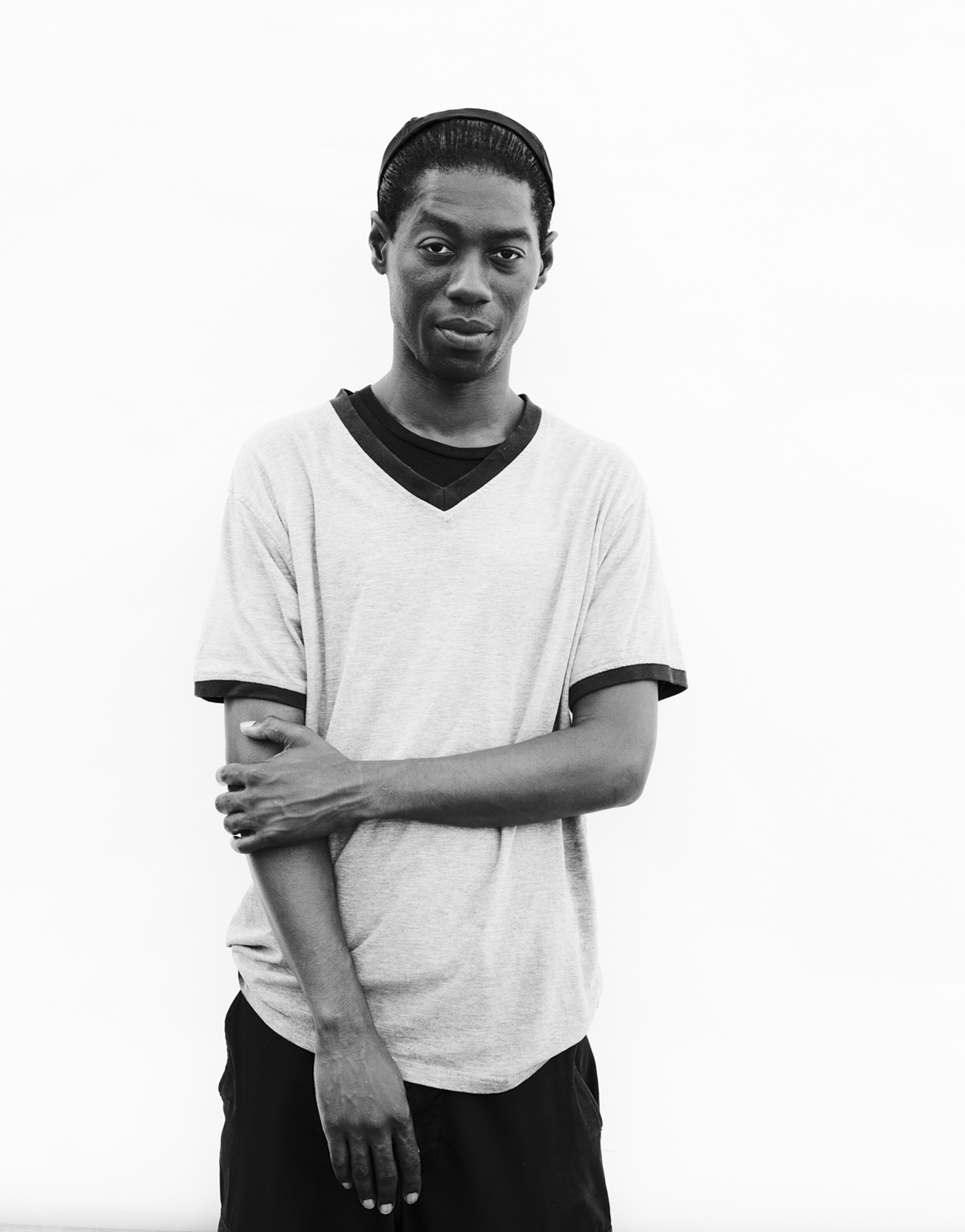 Black and white portrait of a young adult male with short hair, wearing a t-shirt and a head band, standing against a white background.
