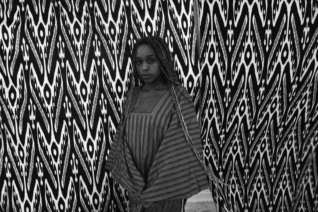Portrait of a woman with braided hair wearing a striped dress, posed in front of a black and white patterned backdrop.