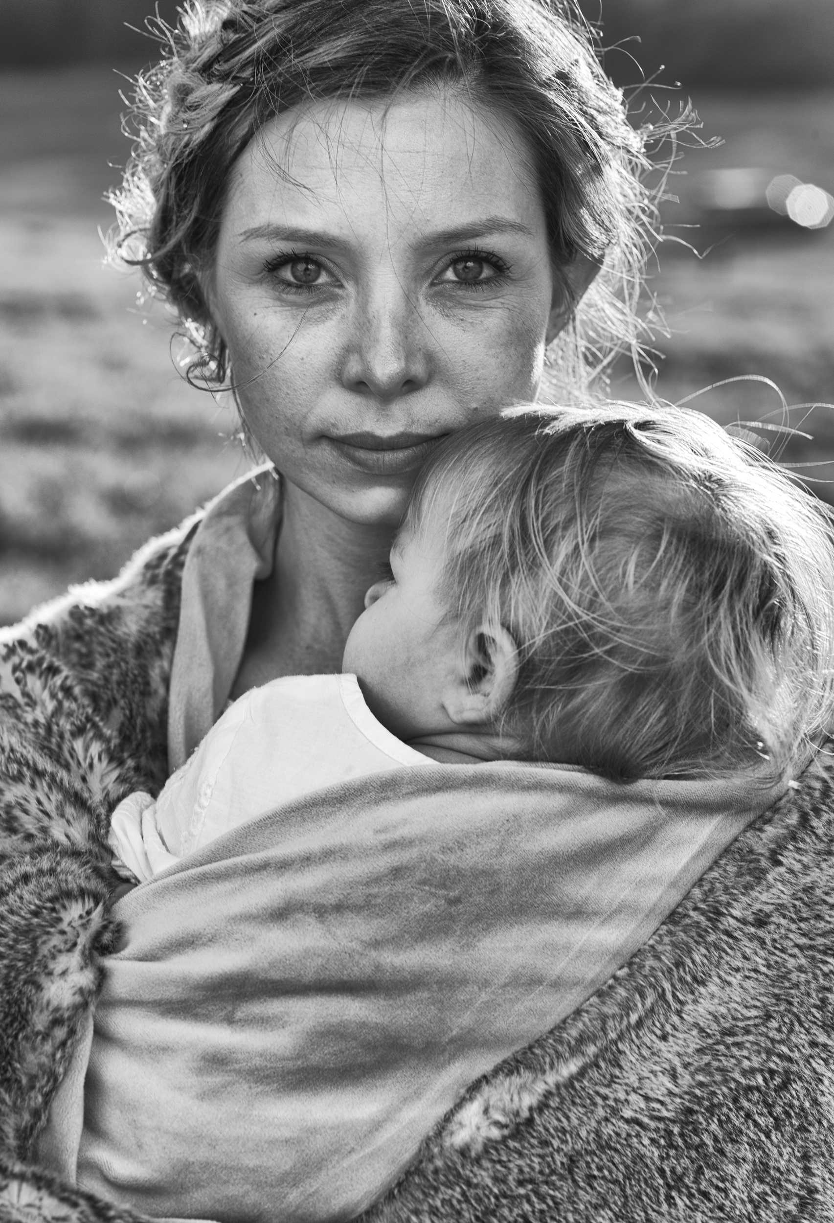 Black and white portrait of a woman holding a baby wrapped in a blanket, both looking peacefully at the camera with a blurred background.