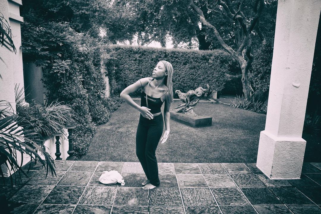 A slender woman with blonde braids performs dance movements barefoot on a tiled patio near a manicured green garden and stone sculpture.
