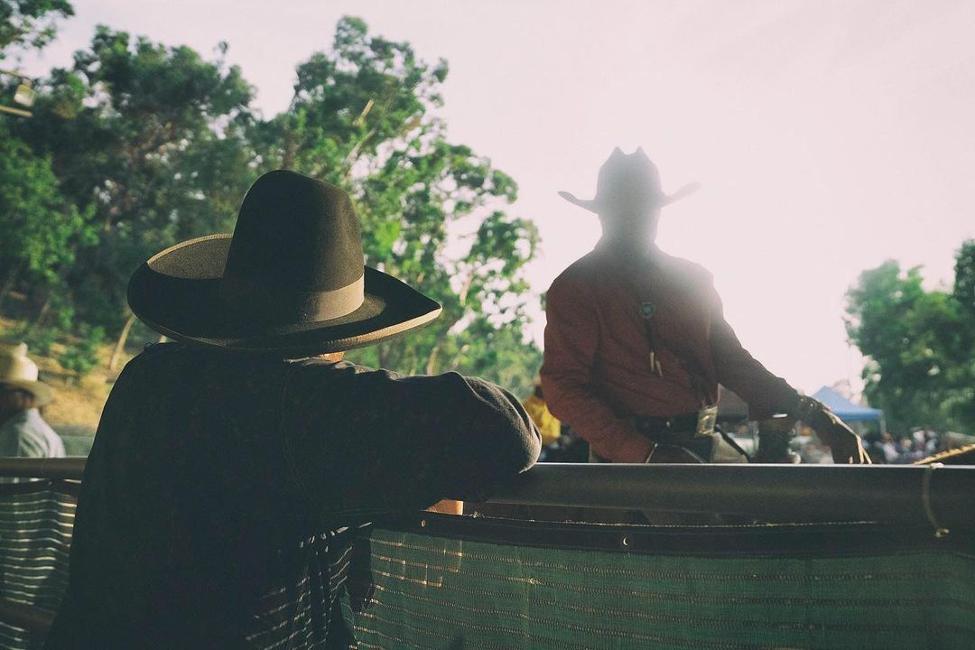 Two people in cowboy hats at an outdoor event, with one person facing away from the camera, wearing a dark coat and hat.