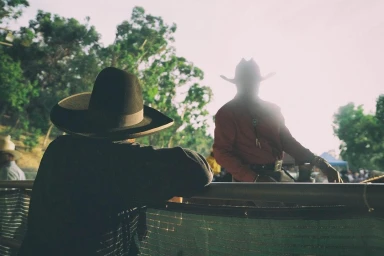 Two people wearing cowboy hats outdoors, one leaning on a railing in the foreground with strong backlighting.