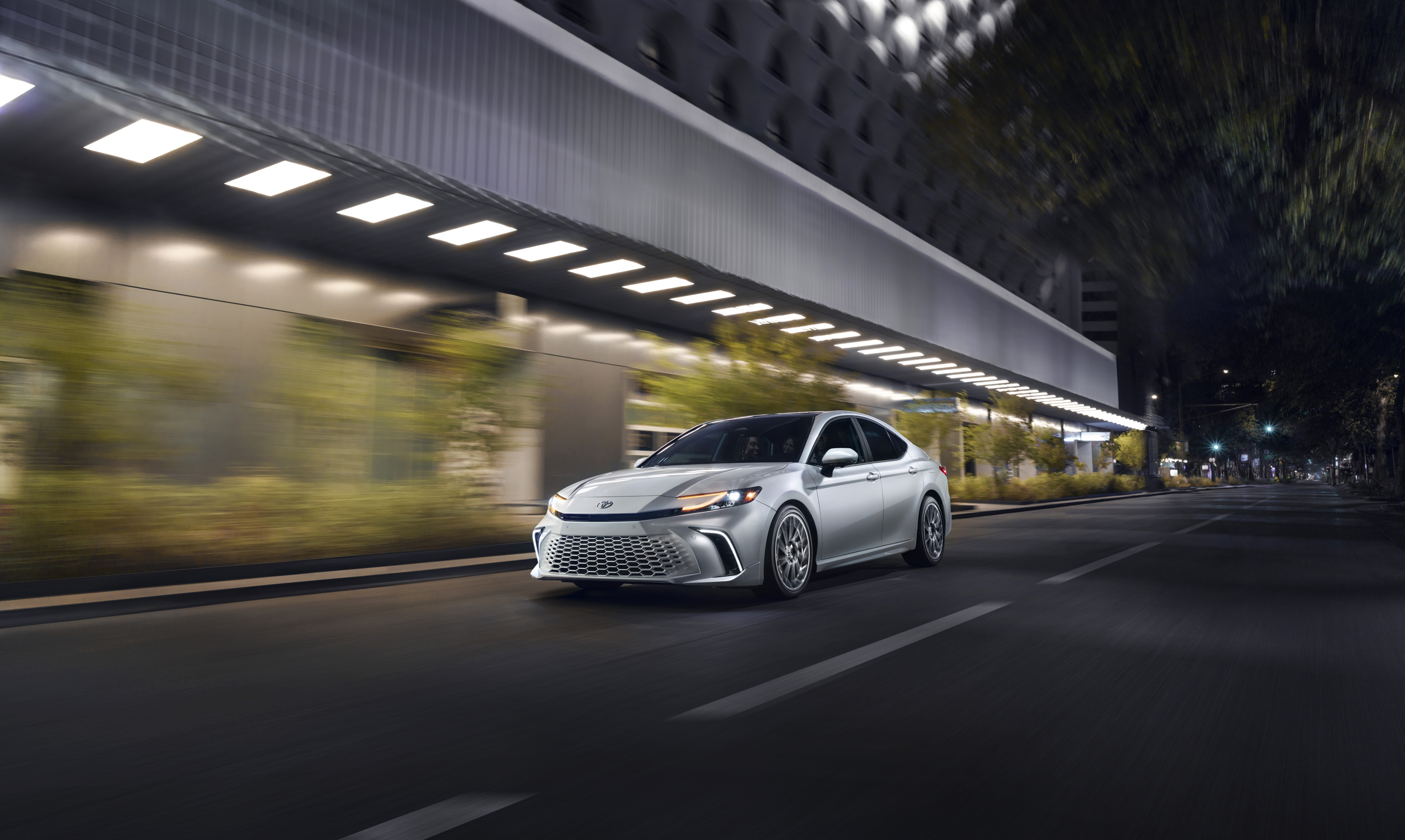 Silver sedan driving on an asphalt road in a city at night with a modern building in the background illuminated by streetlights.