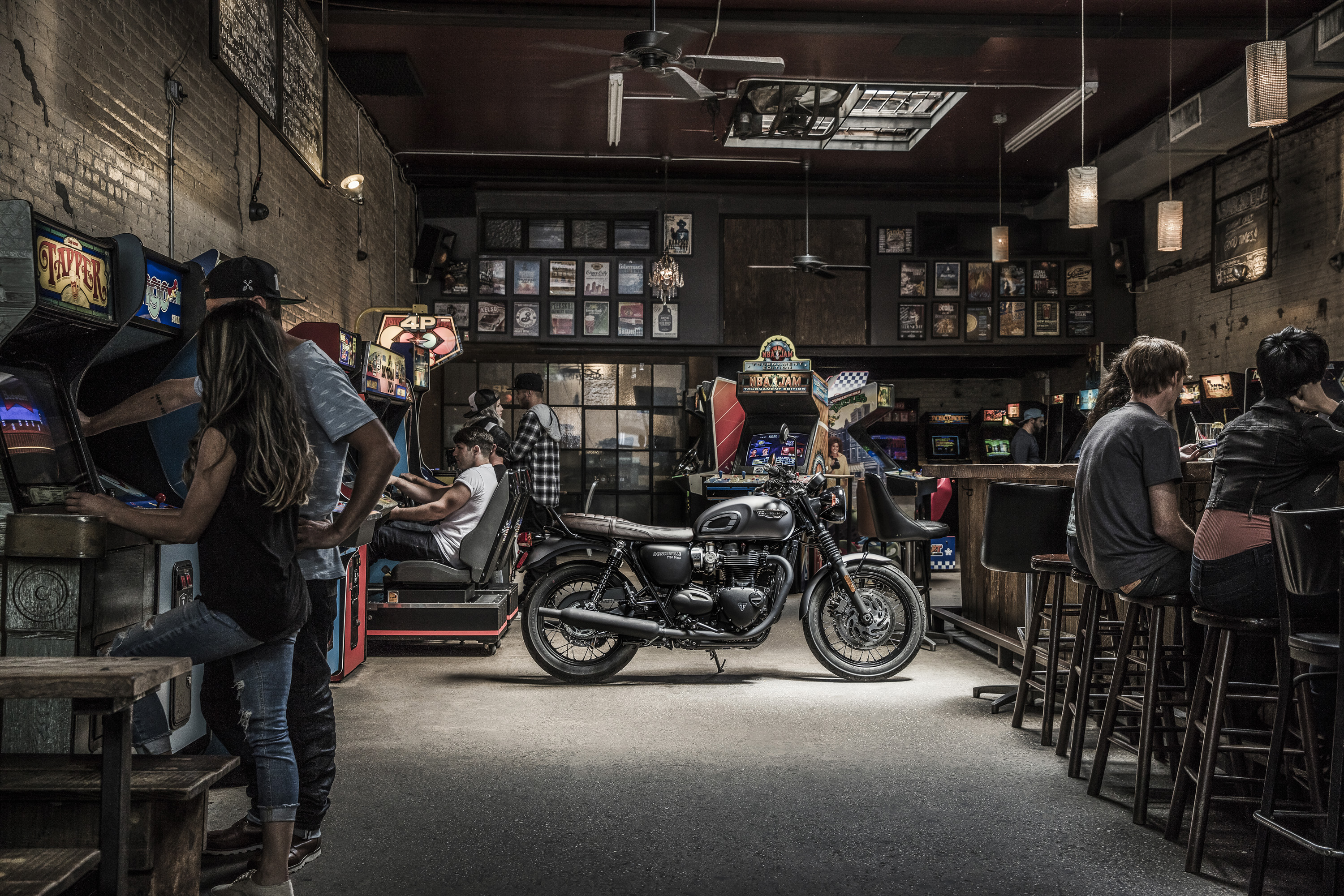 Classic motorcycle parked inside vintage arcade with people playing games and seated at bar; warm lighting and nostalgic atmosphere.