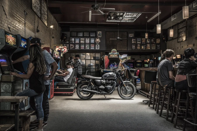 Classic motorcycle parked inside vintage arcade with people playing games and seated at bar; warm lighting and nostalgic atmosphere.