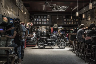 A vintage motorcycle parked inside an arcade bar with patrons playing arcade games and seated at a wooden counter.