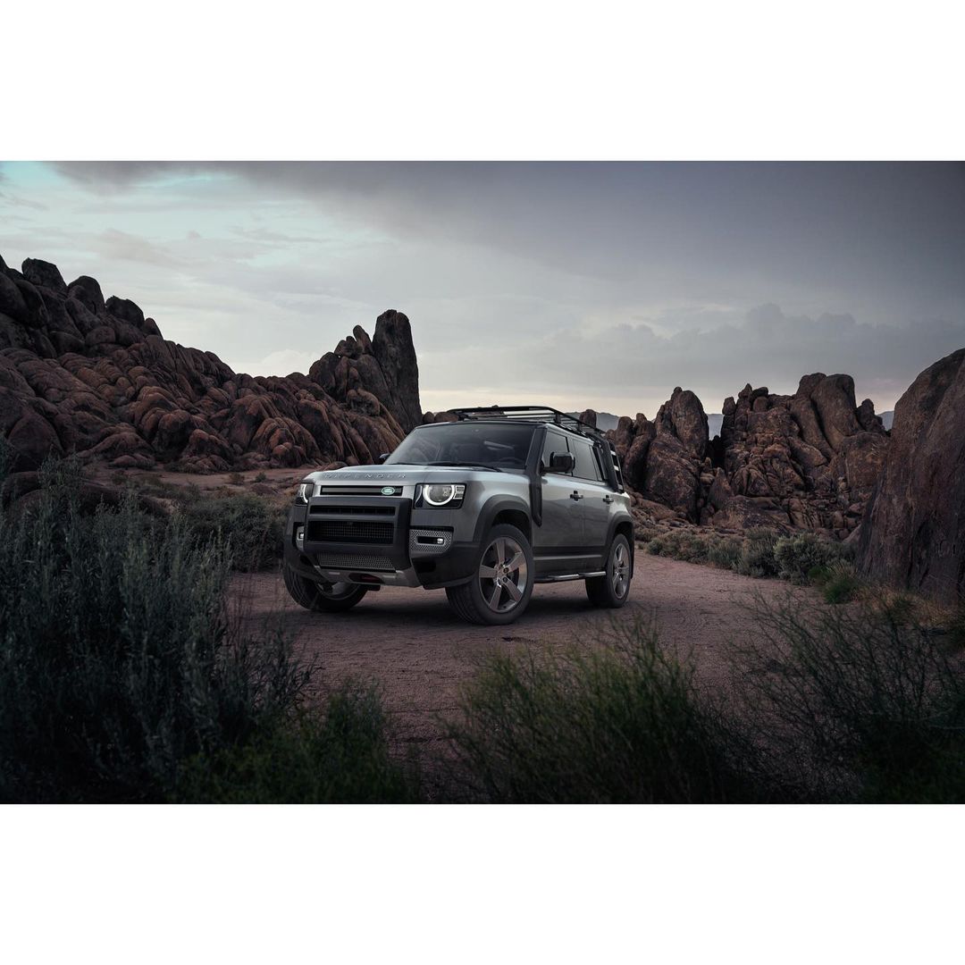Grey Land Rover Defender SUV parked on a dirt road surrounded by desert vegetation and rocky hills under a cloudy sky.
