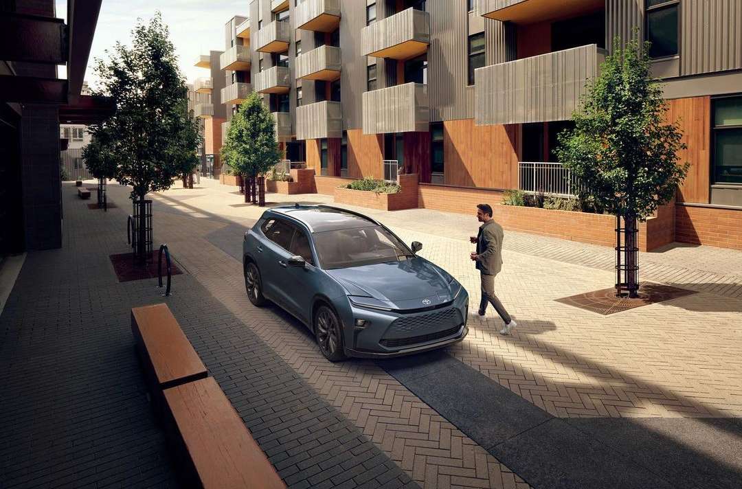 Blue SUV parked on a paved street with a man walking nearby, set against a modern apartment building and tree-lined walkway.