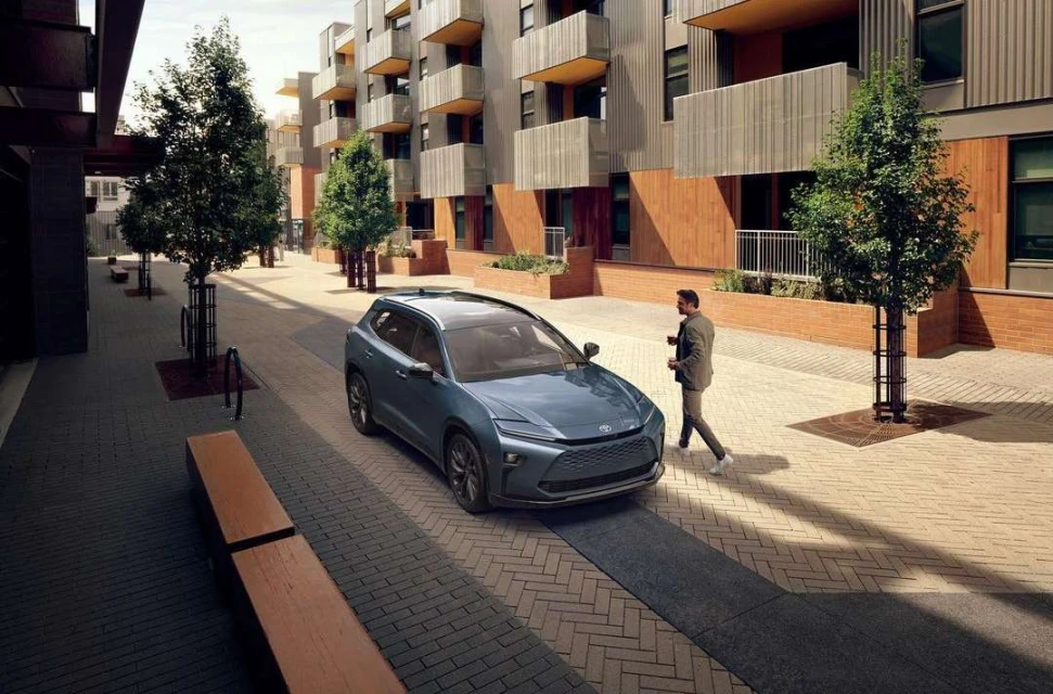 Blue SUV parked on a paved street with a man walking nearby, set against a modern apartment building and tree-lined walkway.