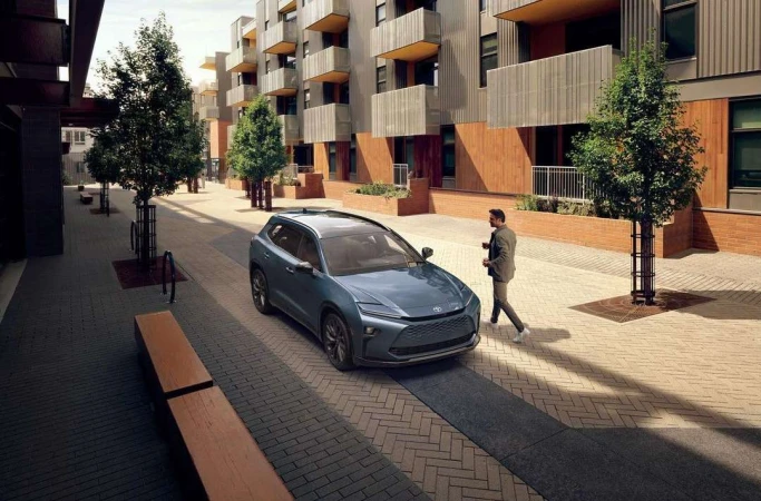 Blue SUV parked on a paved street with a man walking nearby, set against a modern apartment building and tree-lined walkway.