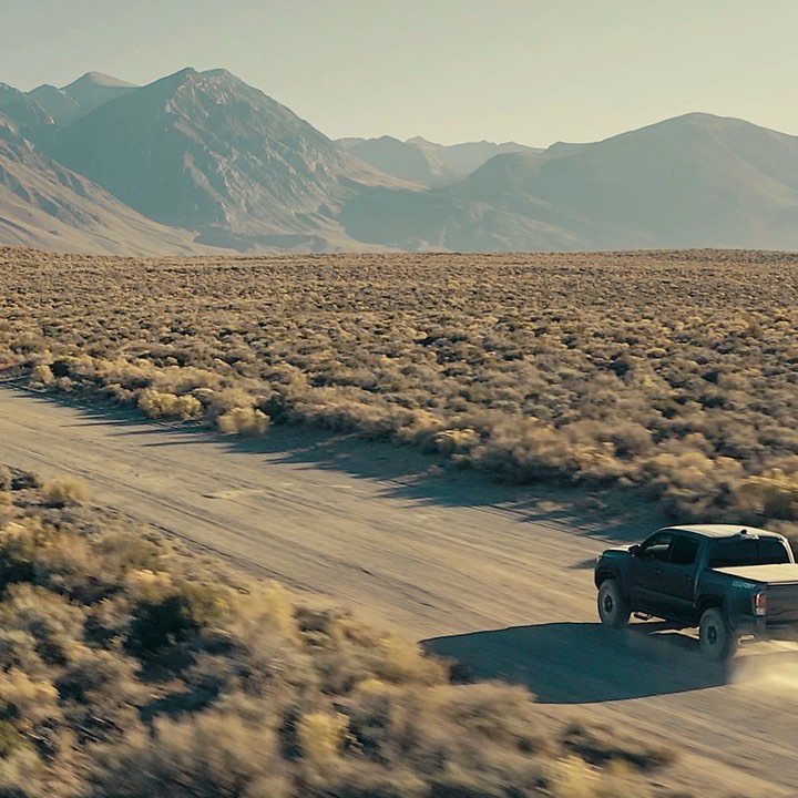 Off-road truck drives through a desert landscape with mountains in the distance on a dusty road, under a bright and clear sky.