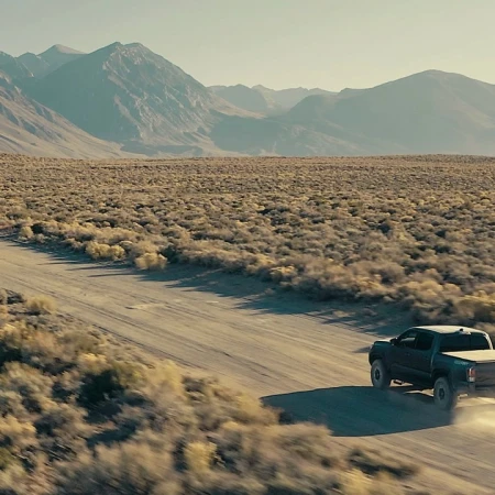 Off-road truck drives through a desert landscape with mountains in the distance on a dusty road, under a bright and clear sky.