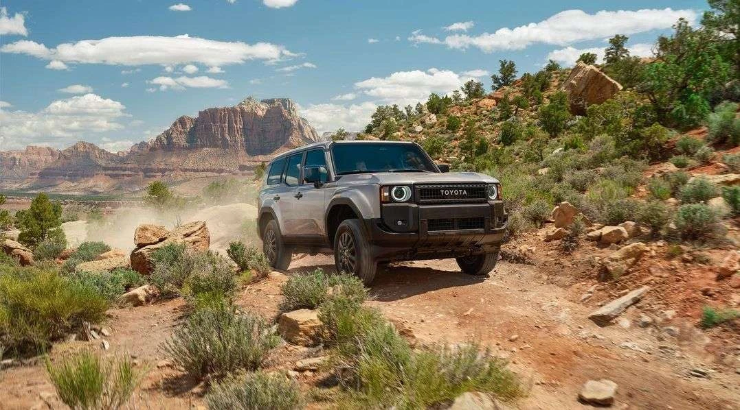 Silver Toyota SUV driving on a dirt road through a desert landscape with mountains and scattered vegetation on a sunny day.
