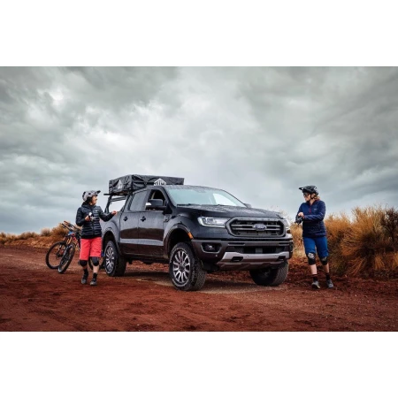 Ford Ranger pickup truck with a roof tent and two mountain bikers on a dirt road under a cloudy sky, showcasing an outdoor lifestyle.