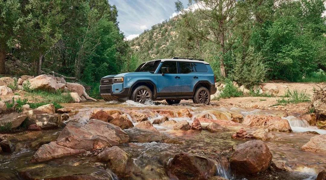 Blue SUV driving through a rocky riverbed surrounded by trees. Automotive scene showcases vehicle in a natural, outdoor environment.