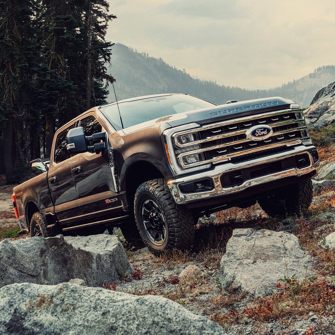 Brown Ford Super Duty truck navigates a rocky, mountainous terrain. Tall pine trees and misty mountains form the scenic backdrop. 