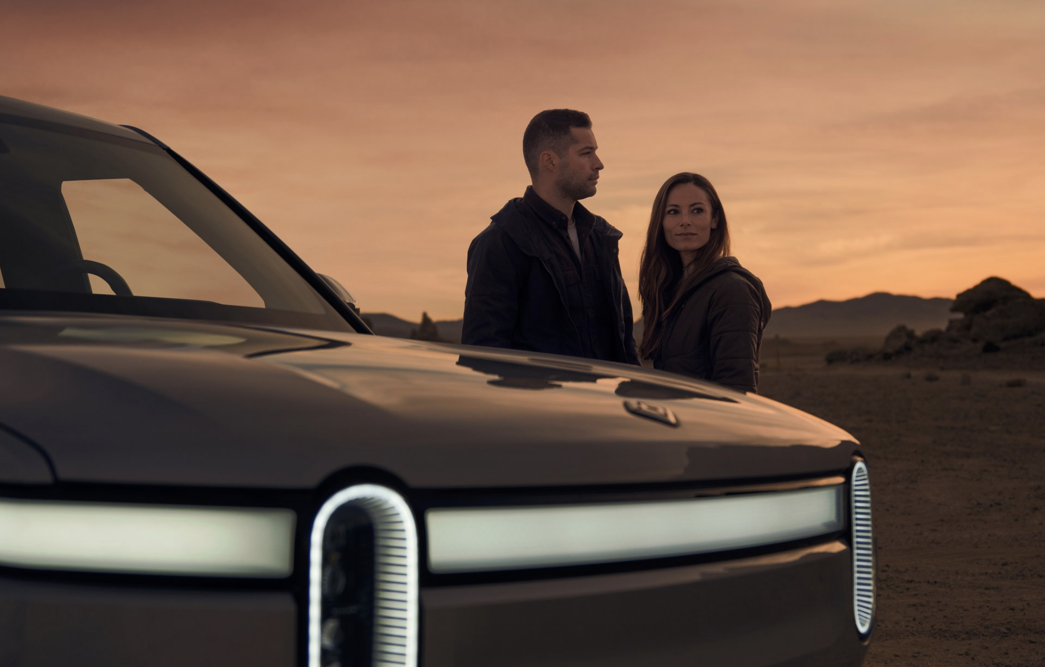 Rivian electric truck in a desert landscape with a man and woman standing behind the vehicle at dusk, displaying modern design.