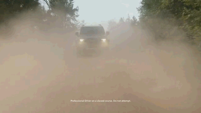 An SUV kicks up dust on a dirt road, shrouded in a cloud of dust and dirt. Trees and sky peek through the background.