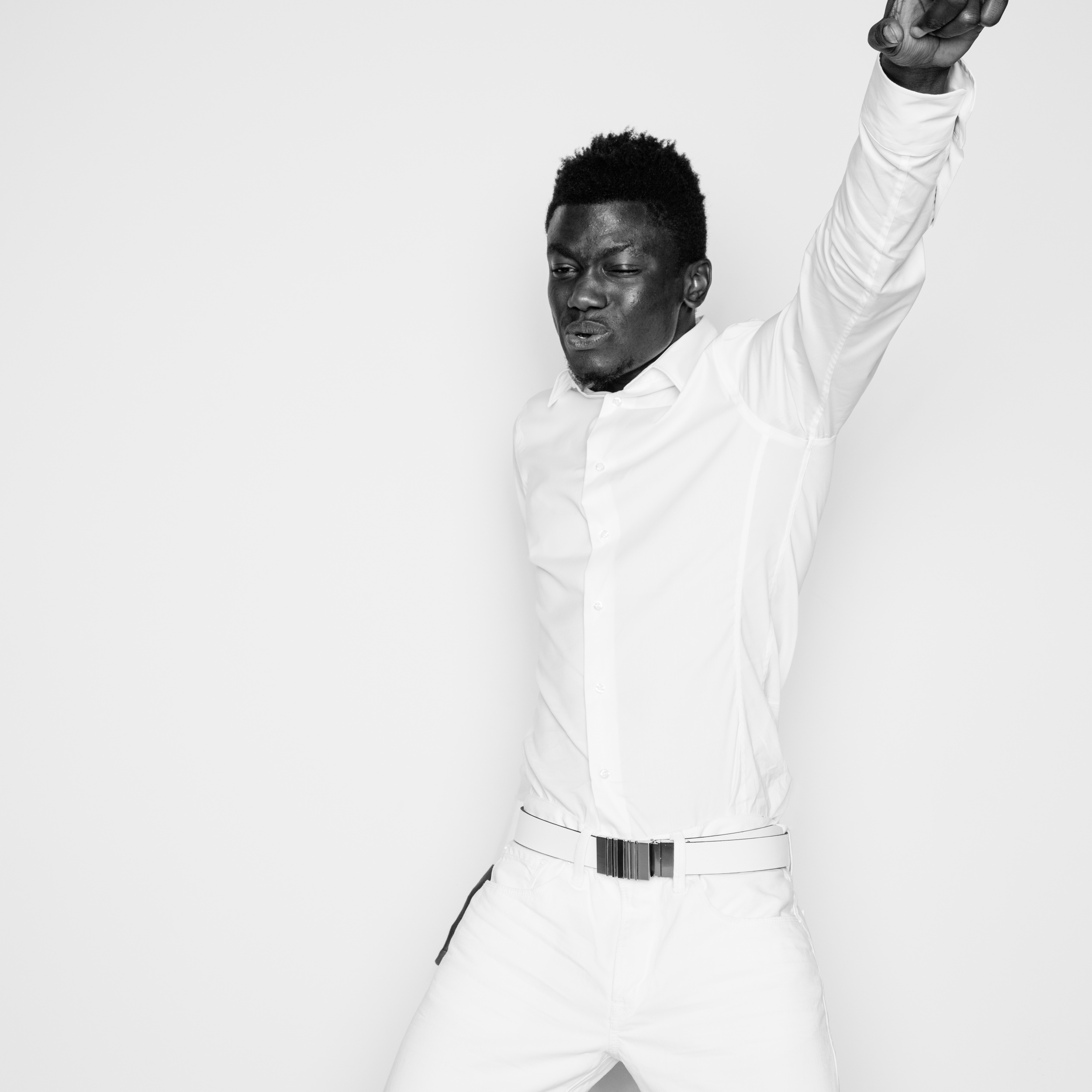Studio shot of an African American man in a white shirt and pants with arm raised against a white backdrop in black and white.