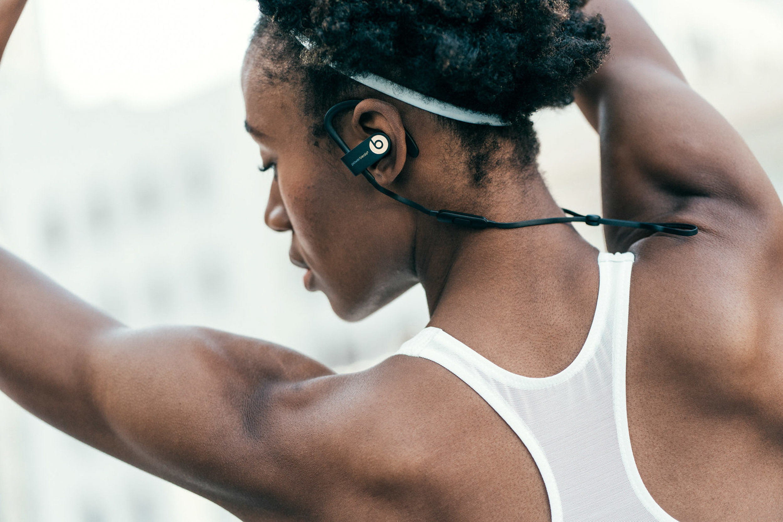 Close-up of athletic adult female stretching her arms behind her head, wearing a white tank top and black Beats headphones outdoors.