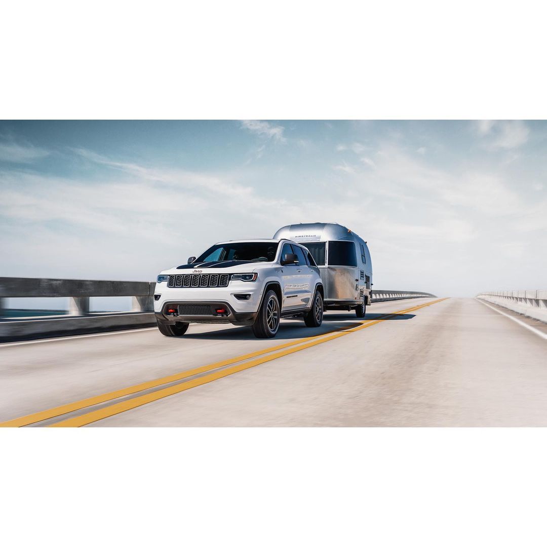 White Jeep Grand Cherokee towing a silver Airstream trailer on a highway under a blue sky. Automotive and travel lifestyle imagery.