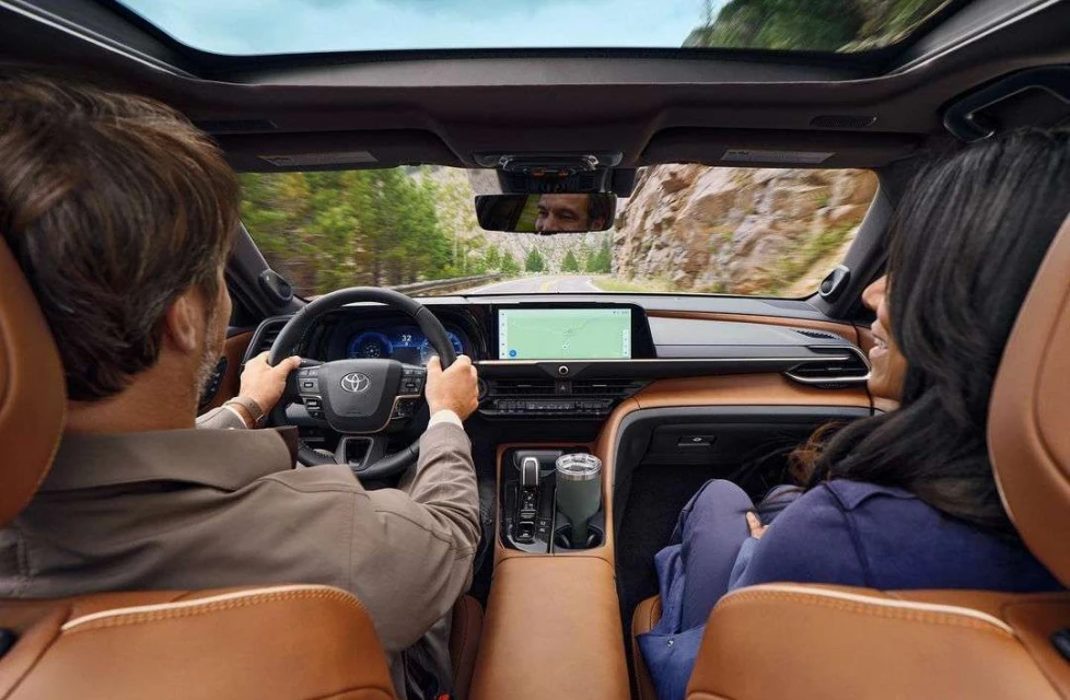 View from inside car with adult male driving, and adult female passenger, passing a rock face on a sunny day. Toyota dashboard visible.