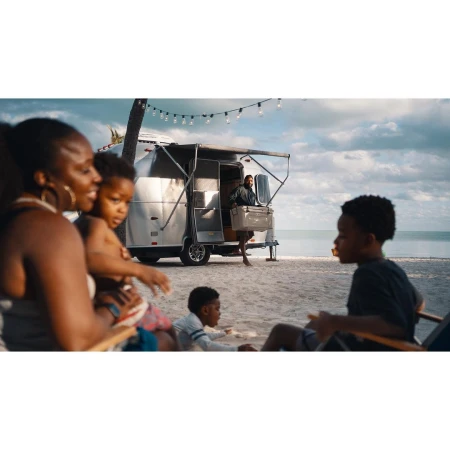 Family enjoys time near a silver trailer on a beach with white sand, blue water, and a palm tree under a partly cloudy sky.