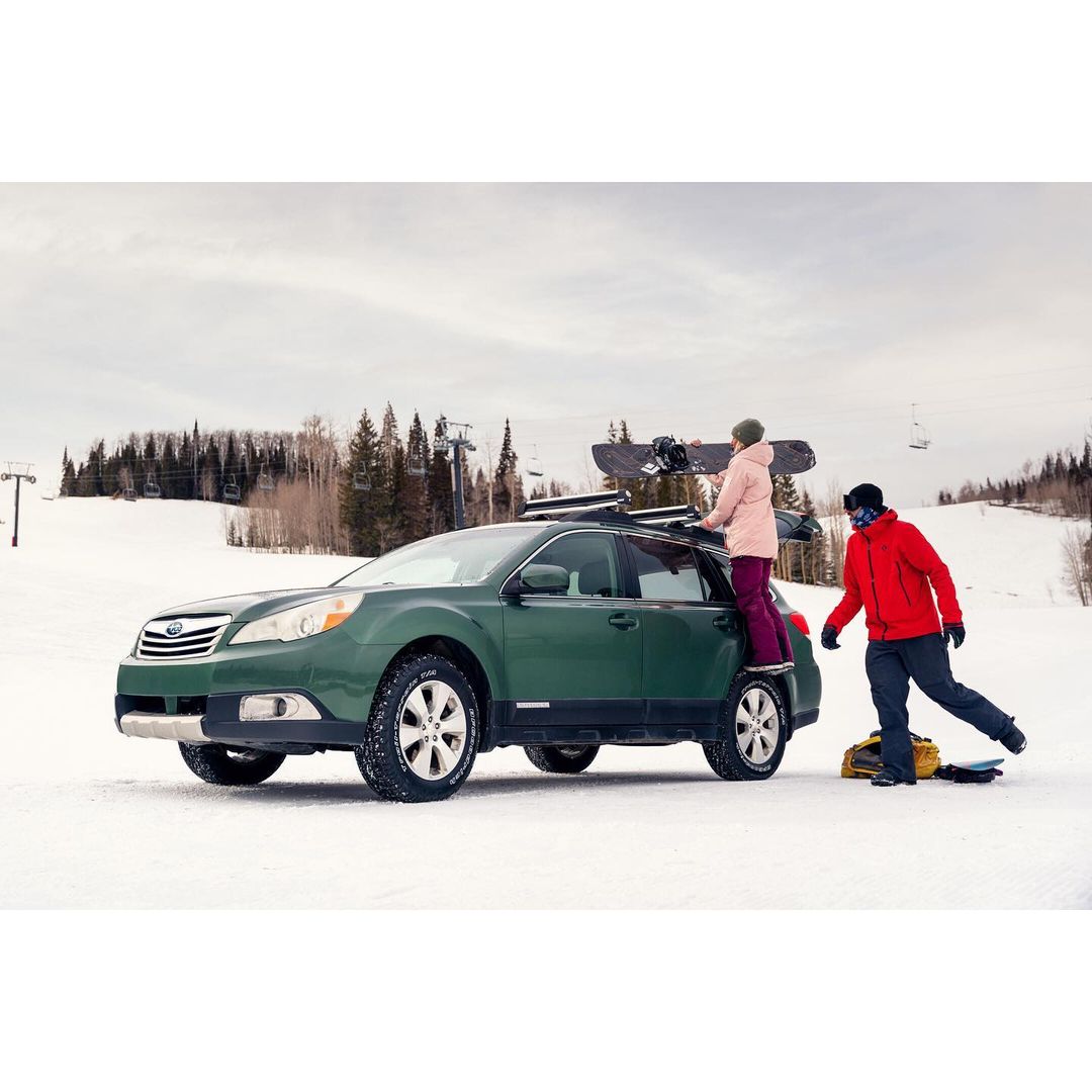 Green Subaru Outback with snowboards on top, parked on snowy hill with two adults nearby, ski resort in background.