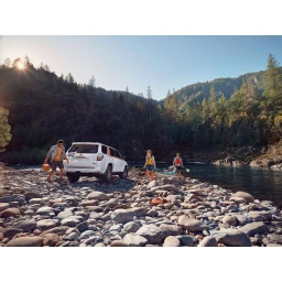People with a white SUV and kayak on a rocky riverbank next to a forest under a bright sunset sky.