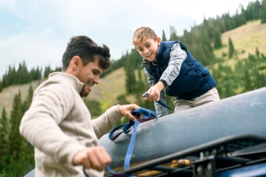 Man and boy securing a canoe on a roof rack outdoors in a mountainous landscape captured by Bax + Towner.