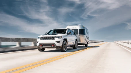 White SUV towing a classic silver travel trailer across a concrete bridge under a partly cloudy sky.