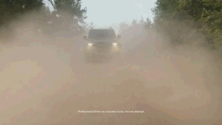 An off-road vehicle driving forward on a dusty dirt path, kicking up thick clouds of brown dust under an overcast sky.