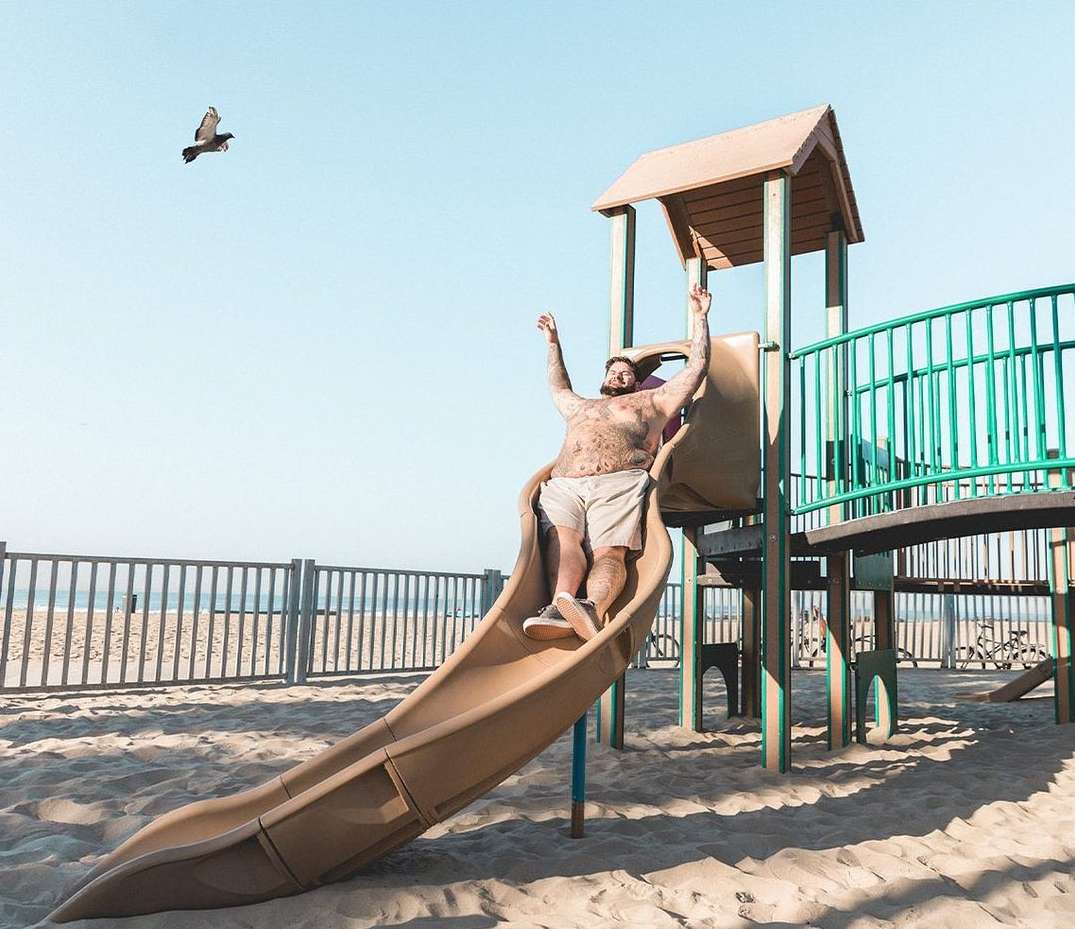 Adult man slides down a tan playground slide at the beach with a bird flying overhead in this outdoor summertime shot.