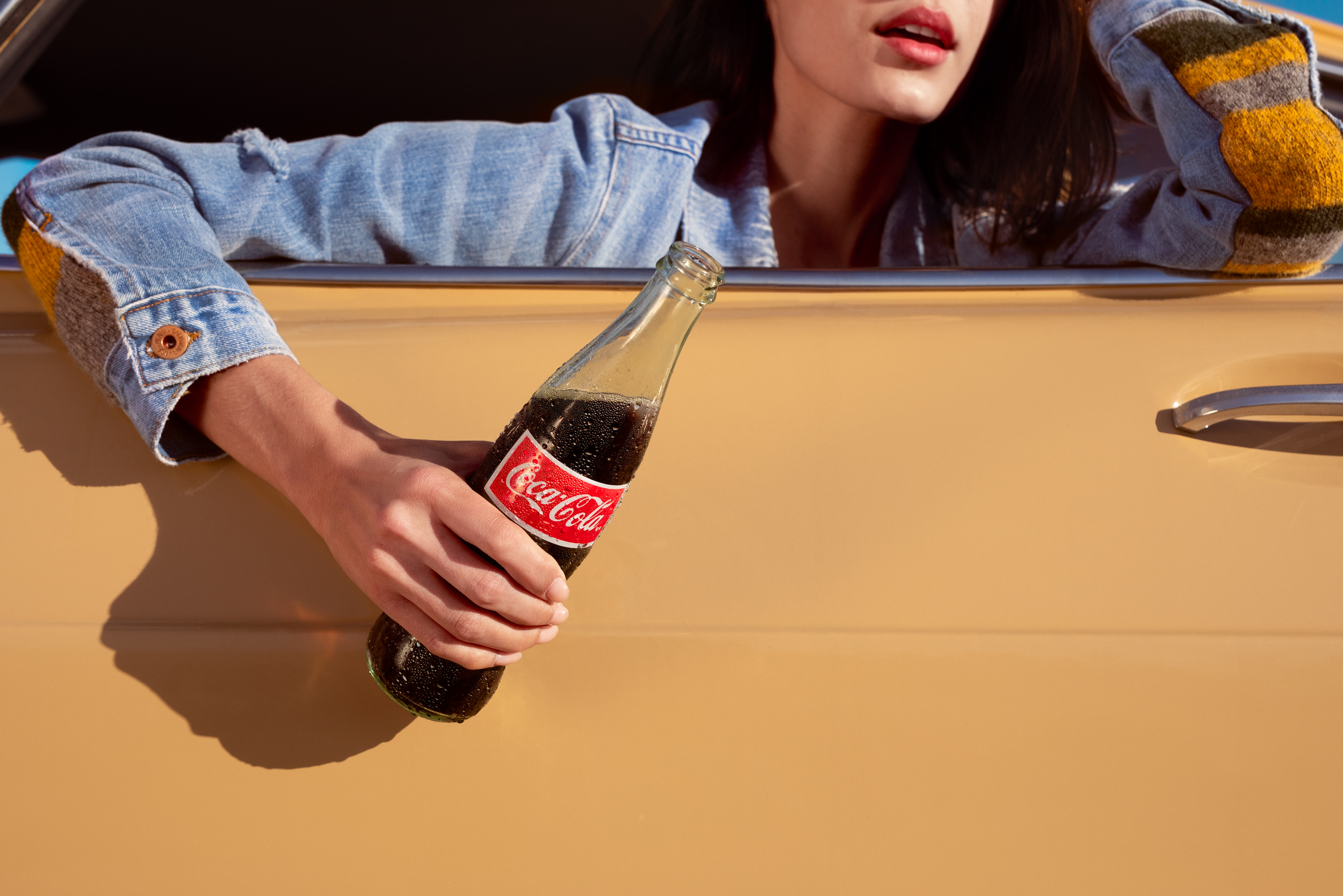 Young woman in a denim jacket holds a glass Coca-Cola bottle while leaning out of the window of a vintage yellow car on a sunny day.