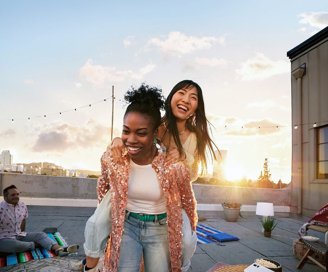 Two young women smiling on a rooftop at sunset. One woman gives the other a piggyback ride in an urban setting during golden hour.