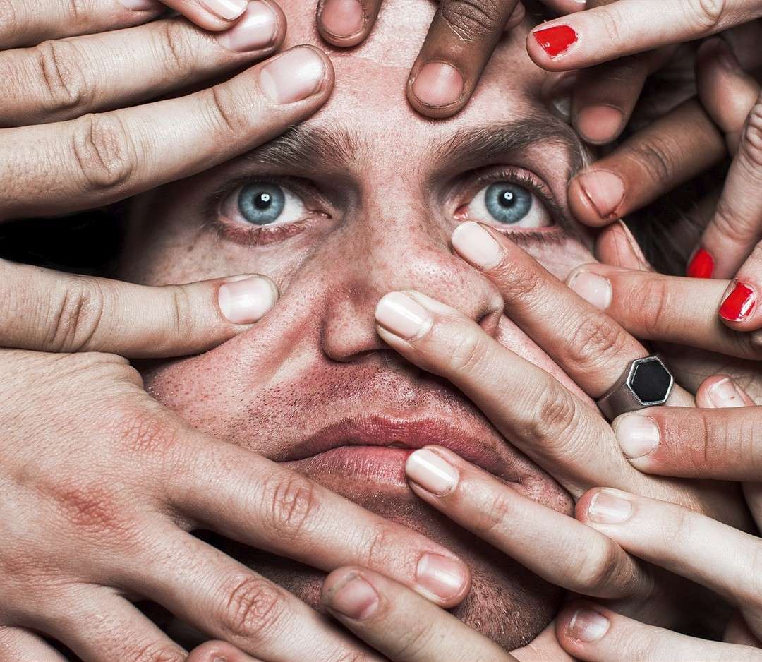 Close-up of a fair-skinned man with blue eyes surrounded by many hands with varied skin tones and painted nails, creating a crowded, intimate portrait.
