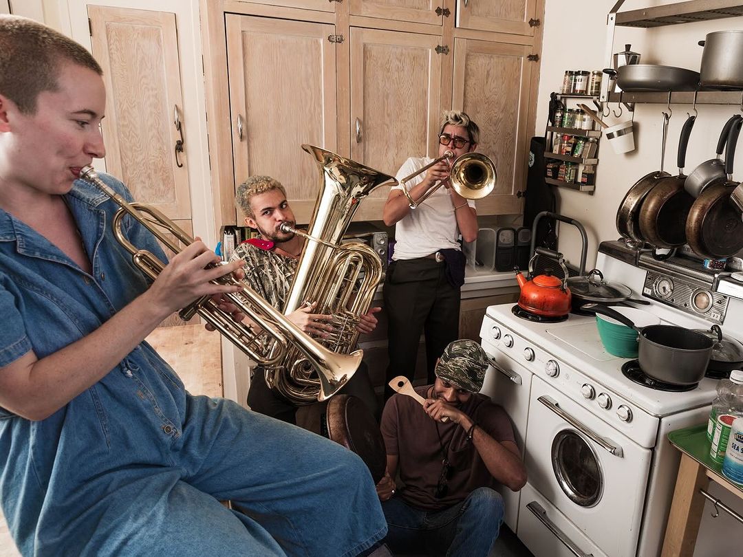 Five musicians play brass instruments, including a trumpet, trombone, and tuba, in a kitchen with white cabinets and cooking equipment.