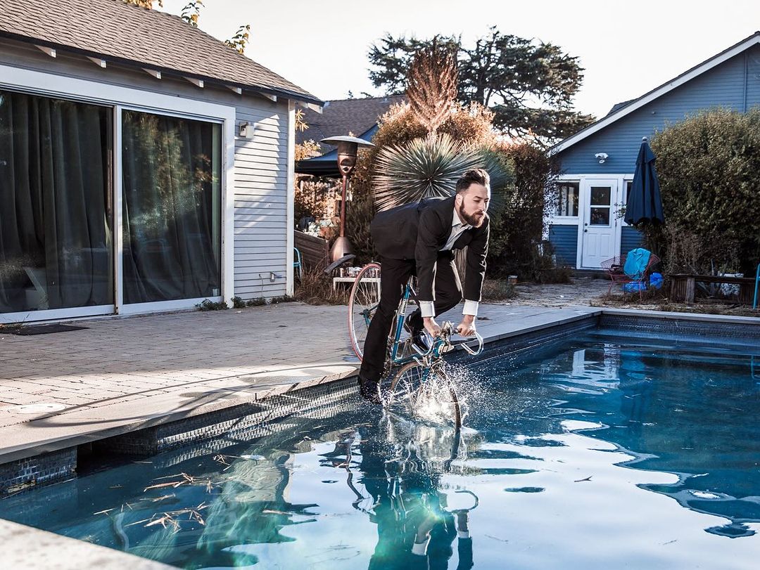 Man in a suit riding a bicycle into a swimming pool in a residential backyard on a sunny day, creating a splash.