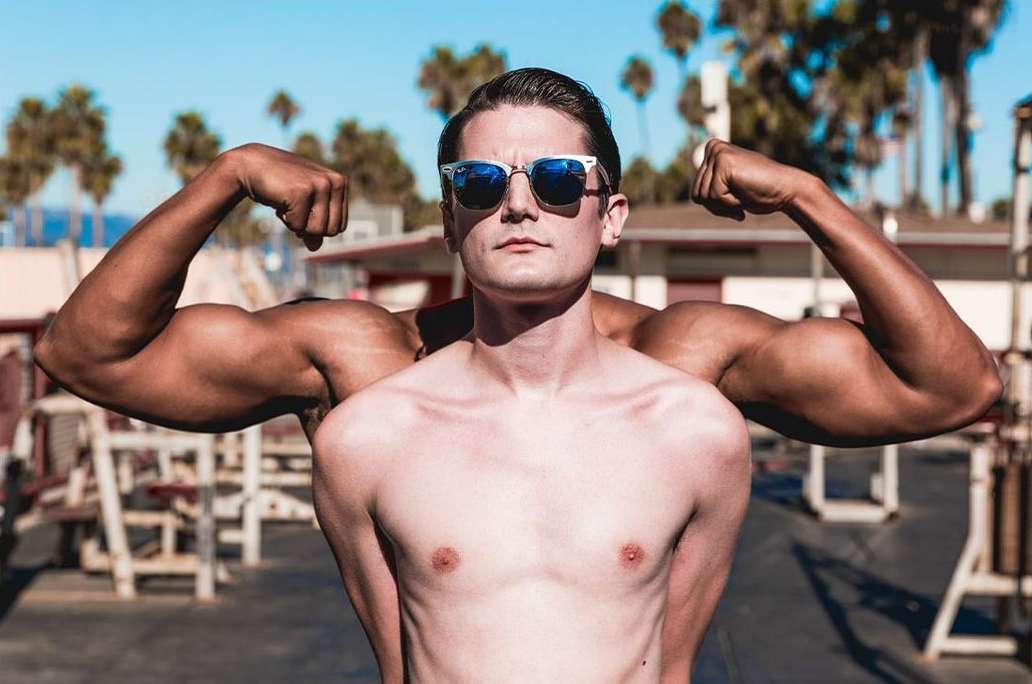 A shirtless man with sunglasses stands in front of another shirtless man flexing his biceps at an outdoor gym on a sunny day.