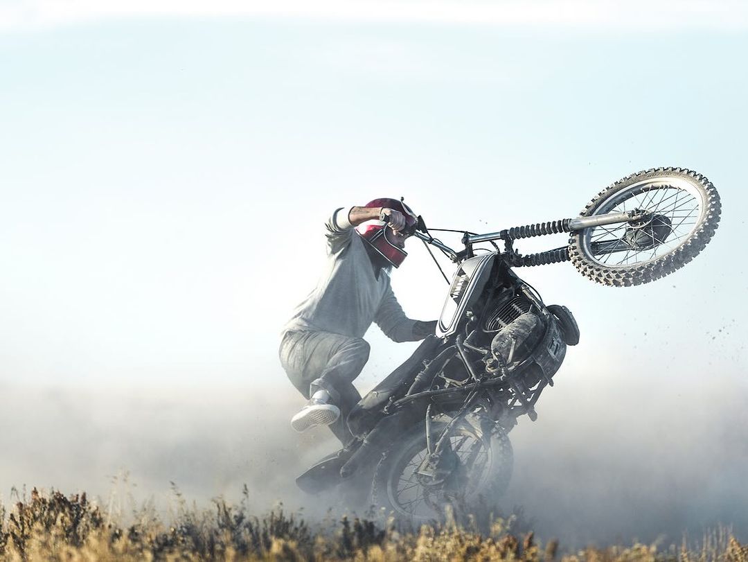 A man on a motorcycle performs a wheelie on a dirt track, kicking up dust, against a bright sky in an outdoor setting.