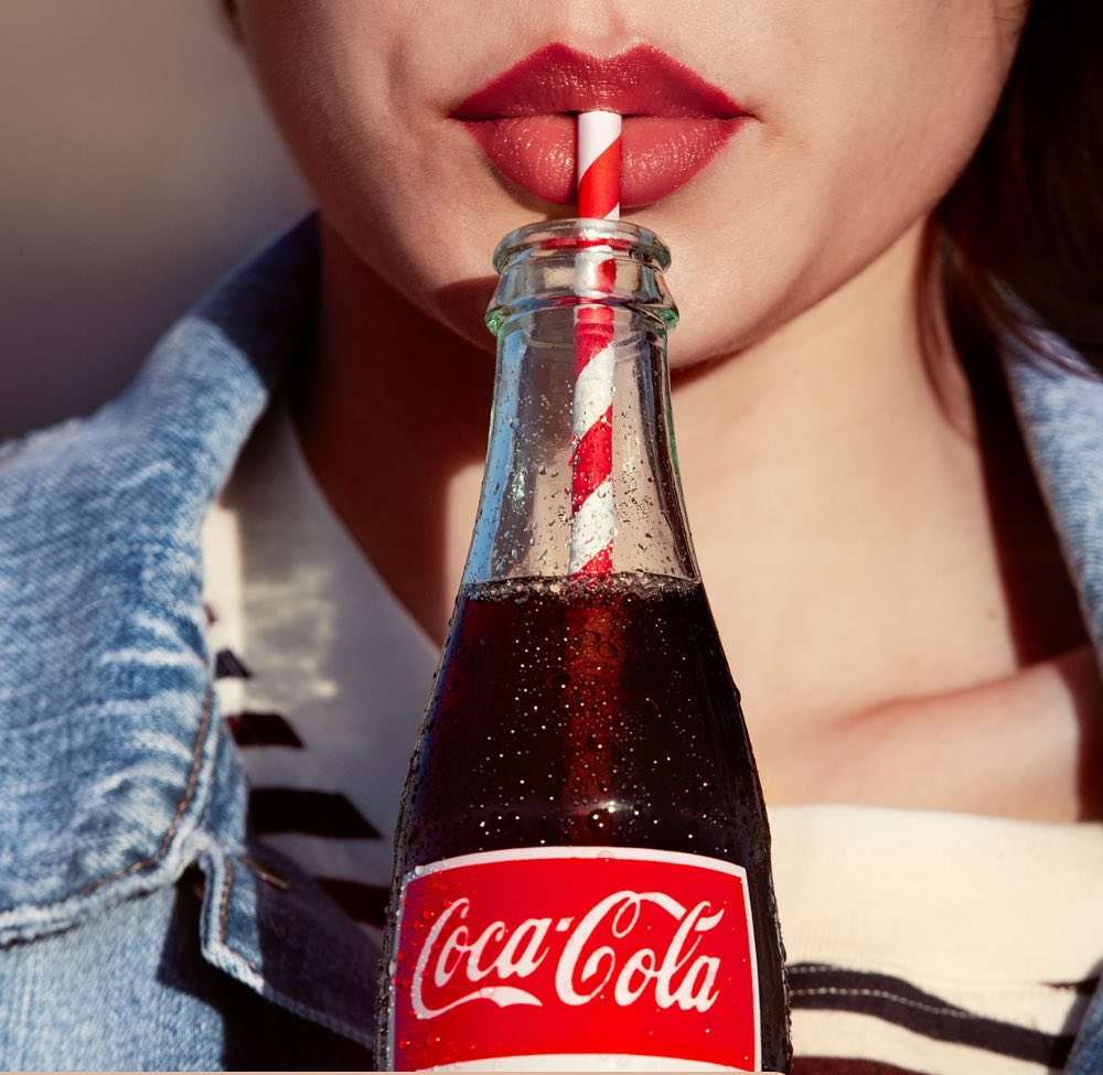 Close-up of a young woman with red lipstick drinking Coca-Cola through a red and white striped straw against a bright background.