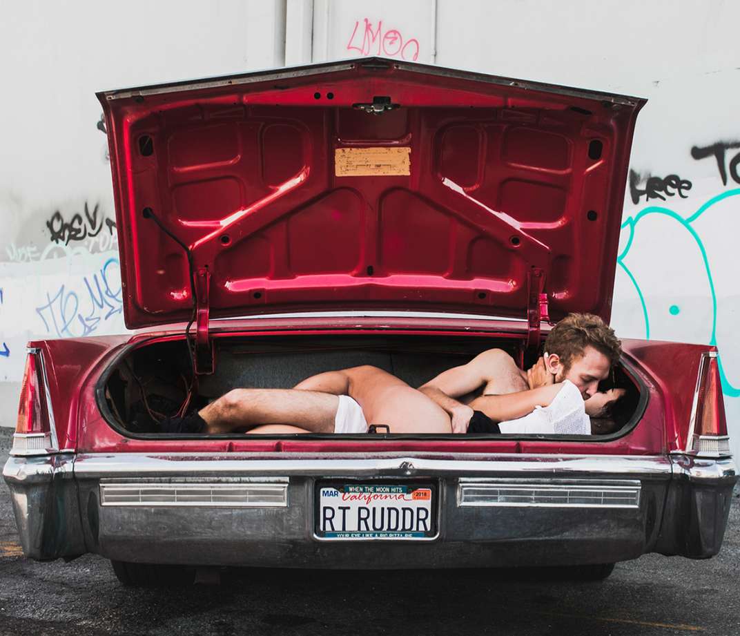 A red vintage car trunk with open lid, inside are two undressed people kissing, with a license plate visible, set against a graffiti wall.